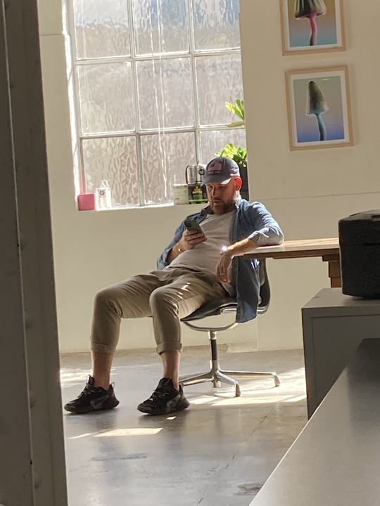 Man sitting in a sunlit room, casually checking his phone while seated at a table, with art on the wall in the background